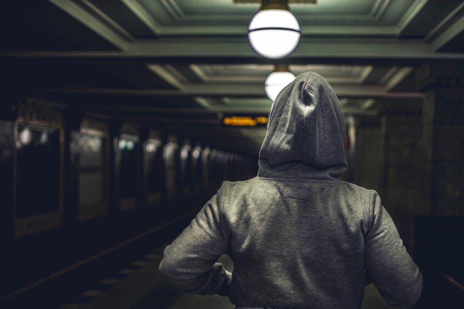 Rear view of a person in a hoodie waiting at a dimly lit subway station platform.
