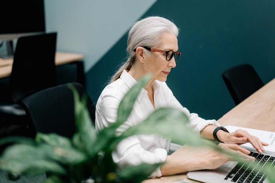 Confident senior woman with glasses working on a laptop in a modern office setting.