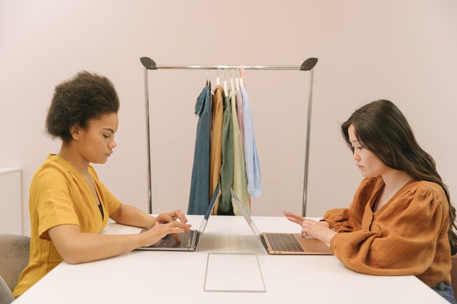 Two women focusing on their laptops in a modern office setting with a clothes rack in the background.
