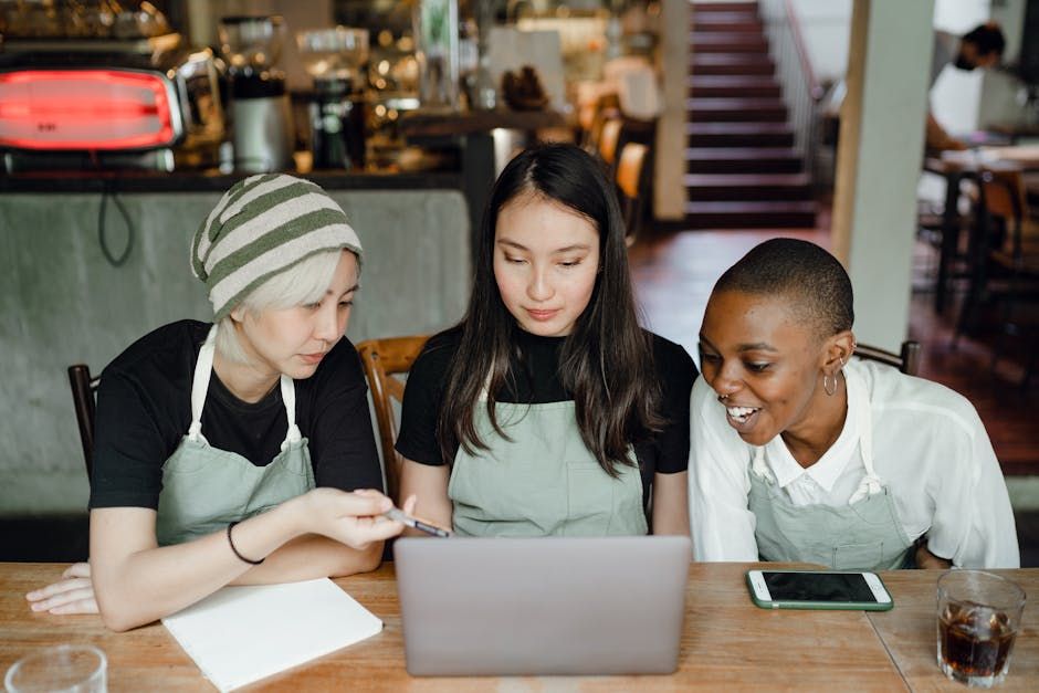 Three café workers in aprons collaborate cheerfully using a laptop indoors.